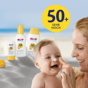 Eine Frau trägt Sonnencreme auf das Gesicht eines Babys auf, im Hintergrund ist ein Strand zu sehen. Die Sonnencreme ist mit "Hipp BabySunFit" beschriftet und hat einen Lichtschutzfaktor von 50+.