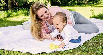 Ein freudiger Moment zwischen einer Frau und einem Baby, wobei die Frau auf einer Decke in einem Park liegt und beide in eine spielerische Aktivität vertieft sind.