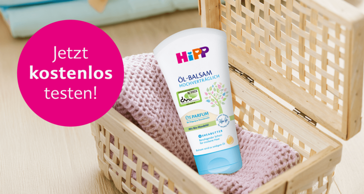 Eine Tube eines Babypflegeprodukts mit der Aufschrift "HiPP" und dem Text "OL-BALSAM HOCHVERTRAGLICH" sowie einem "0% PARFUM"-Etikett. Das Produkt befindet sich in einem geflochtenen Korb mit einem rosa Tuch.