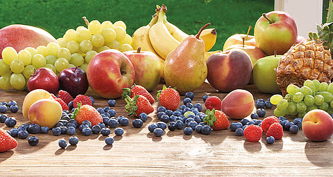 Eine Vielzahl von frischen Früchten und Beeren, die auf einer hölzernen Oberfläche angeordnet sind, darunter Äpfel, Bananen, Trauben, Birnen, Erdbeeren, Heidelbeeren und eine Ananas.