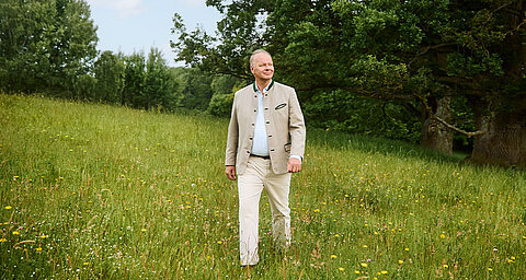 Ein Mann, der in einer beigen Jacke und einer weißen Hose durch eine saftig grüne, mit gelben Blumen übersäte Wiese geht.