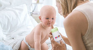 Ein Baby, das neugierig zu sein scheint, spielt mit einer Flasche mit der Aufschrift "Hipp Milk Lotion", die von einer Frau gehalten wird.