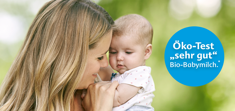 Eine Frau, die ein Baby an sich drückt, mit einem blauen kreisförmigen Logo im Vordergrund, auf dem "Öko-Test Sieger Bio-Babymilch" steht.