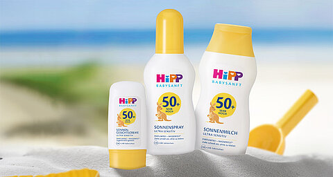 Drei Sonnenschutzprodukte der Marke "HiPP" mit unterschiedlichem Lichtschutzfaktor, platziert an einem Sandstrand mit einem klaren blauen Himmel im Hintergrund.