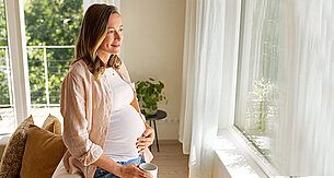 Eine vermutlich schwangere Frau hält eine Tasse in der Hand und blickt aus einem Fenster mit Blick ins Grüne.