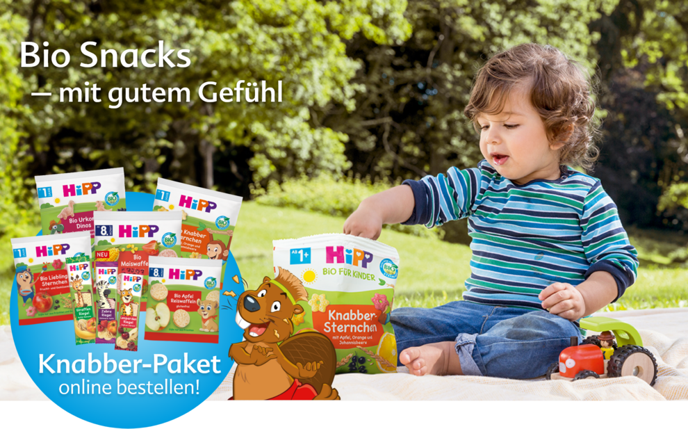 Ein kleines Kind sitzt im Freien und hält eine "Hipp Bio Fur Kinder"-Snackpackung in der Hand, während im Hintergrund verschiedene andere "Hipp Bio Fur Kinder"-Packungen ausgestellt sind.