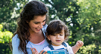 Eine junge Frau und ein kleines Kind im Freien, wobei die Frau dem Kind ein Buch mit dem Titel "BLUBB" vorliest.