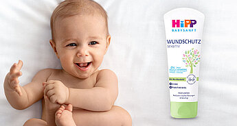 Ein Baby mit freudigem Gesichtsausdruck, das auf einer weißen Fläche liegt, neben einer Tube "WUNDSCHUTZ SENSITIV"-Creme, die mit "Mit Bio-Mandelblend", "21% Panthenol" und "Parfumfrei" gekennzeichnet ist.