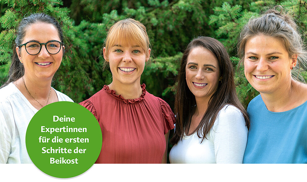Vier Frauen in unterschiedlicher Kleidung posieren im Freien vor einer grünen Kulisse. Ein kreisförmiges Abzeichen in der unteren rechten Ecke enthält einen deutschen Text, der übersetzt "Experten auf dem Gebiet der Beikost" bedeutet.
