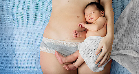 Eine Mutter hält ihr neugeborenes Baby, das in ein weißes Tuch gewickelt ist, während die Mutter hellblaue Unterwäsche mit Spitzenverzierung trägt.