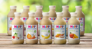 Eine Reihe von Smoothies der Marke "Hipp", die jeweils verschiedene Obst- und Gemüsesorten enthalten und auf einer hölzernen Oberfläche mit einem verschwommenen grünen Hintergrund präsentiert werden.