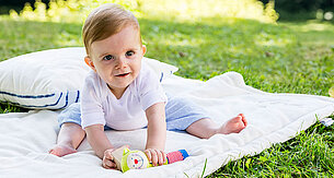 Ein Baby sitzt auf einer Decke im Freien und hält ein buntes Spielzeug.