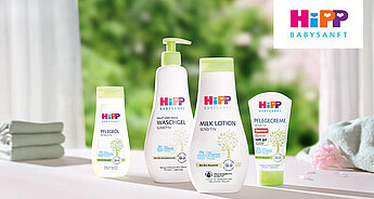 Eine Reihe von Babypflegeprodukten von Hipp Babysanft, darunter eine Haarwäsche, eine Babylotion und eine Pfandcreme, vor einem Hintergrund aus Grünpflanzen und einem gefalteten Handtuch.