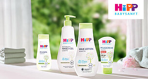 Eine Reihe von Babypflegeprodukten von Hipp Babysanft, darunter eine Haarwäsche, eine Babylotion und eine Pfandcreme, vor einem Hintergrund aus Grünpflanzen und einem gefalteten Handtuch.