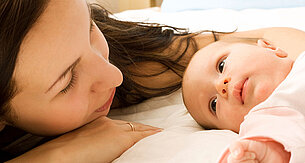 Ein zärtlicher Moment zwischen einer Mutter und ihrem Baby, in dem die Mutter ihr Gesicht sanft neben das des Babys legt und beide einen heiteren und intimen Moment zu teilen scheinen.