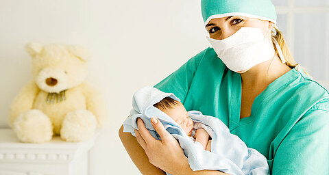 Eine medizinische Fachkraft in grüner Kleidung hält ein neugeborenes Baby, mit einem Teddybär im Hintergrund.