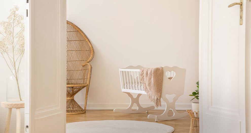 Ein ruhiges Kinderzimmer mit einem weißen Kinderbett, einem Korbsessel und einer hohen Glasvase mit getrockneten Pflanzen.