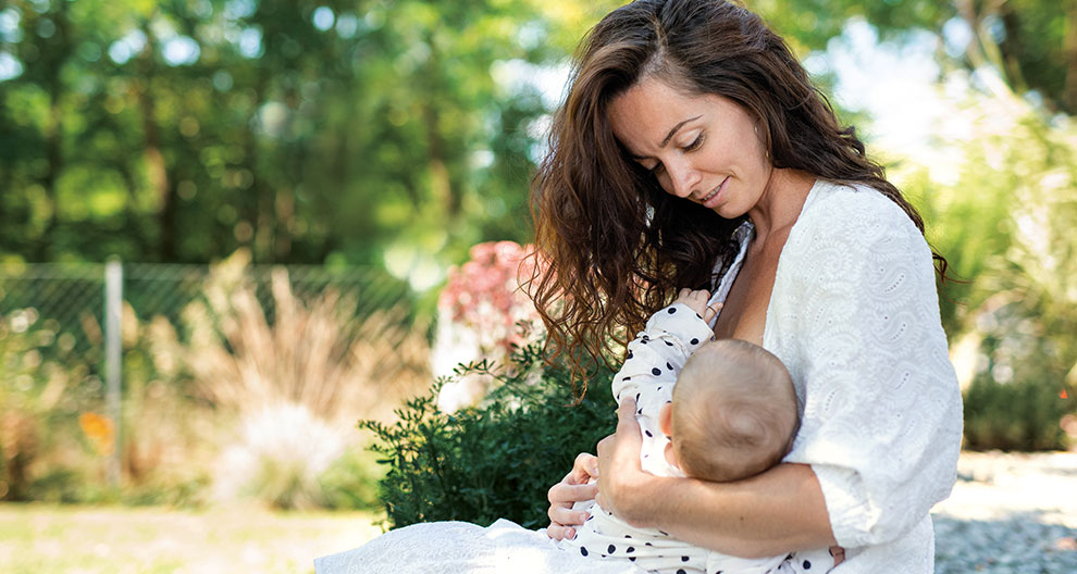 Eine Frau mit langen braunen Haaren, die sanft ein Baby an ihre Brust drückt, im Freien in einer Gartenanlage.