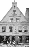 Ein historisches Gebäude mit mehreren Fenstern, einem Schild mit der Aufschrift "Friedrich Krug" und mehreren Personen, die davor stehen.