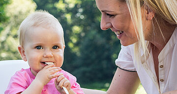 Ein kleines, rosa gekleidetes Baby kaut auf einem weißen Gegenstand, während eine Frau, die vermutlich die Mutter des Babys ist, es anlächelt.