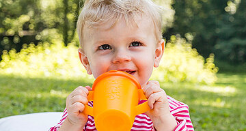 Ein kleines Kind, vermutlich ein Kleinkind, hält eine orangefarbene Schnabeltasse im Freien.