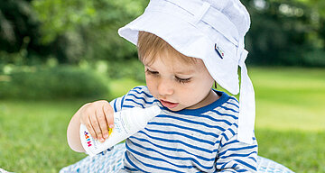 Ein kleines Kind mit weißem Hut und gestreiftem Hemd, das Sonnencreme aus einer weißen Flasche aufträgt.