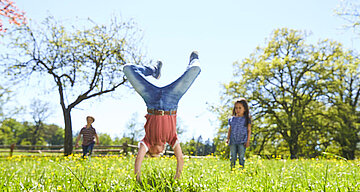 Ein fröhlicher Moment im Freien mit einem Kind, das einen Handstand inmitten eines Blumenfeldes macht, während zwei andere Kinder zusehen.