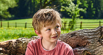 Ein kleiner Junge mit kurzen, zerzausten Haaren und einem rosa Hemd sitzt vor einer grünen Wiese mit einem Holzzaun im Hintergrund.