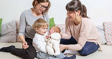 Zwei Frauen, eine mit einem Baby im Arm, die andere mit einem Kleinkind, beide auf dem Boden sitzend, mit einer weißen Wand und grünen Polstern im Hintergrund.