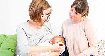 Zwei Frauen, eine davon mit einem Baby in der Hand, in einem Raum mit weißem Hintergrund, mit einem grünen Kissen und einem blumengemusterten Kissen im Hintergrund.