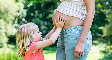 Ein junges Mädchen mit blondem Haar, das sanft den Bauch einer schwangeren Frau berührt, vor einem Hintergrund aus Grün.