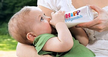 Ein Baby trinkt Milch aus einer Flasche mit der Aufschrift "HiPP Folgemilch", die von einer Person gehalten wird.