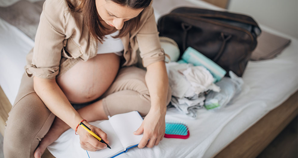 Schwangere Frau sitzt auf einem Bett und schreibt in ein Notizbuch, neben ihr liegen Babykleidung und eine Tasche.