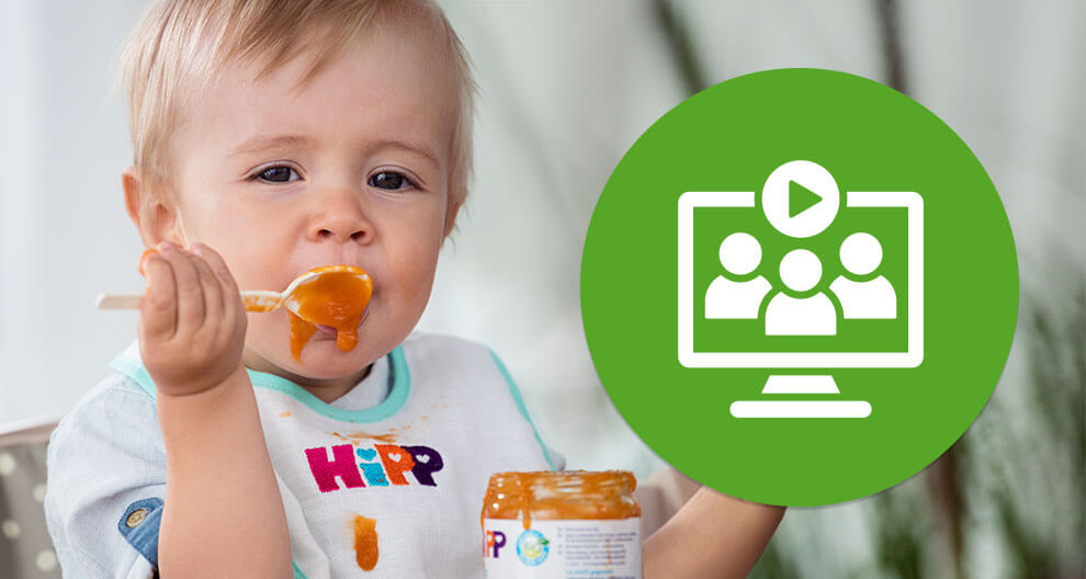 Ein Baby isst einen Löffel mit orangefarbenem Essen. Auf der rechten Seite ist ein grünes, kreisförmiges Symbol zu sehen, das einen Computerbildschirm mit drei Silhouetten darstellt.